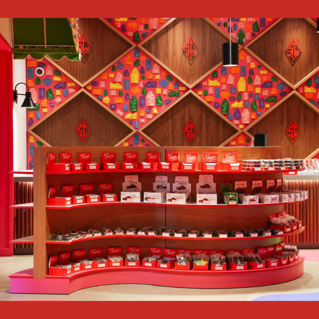 Sarris Candy Floor Display | BAW Plastics Inc. | BAW Plastics Inc. Curved red display shelves stocked with red-labeled chocolate boxes in a colorful shop interior.