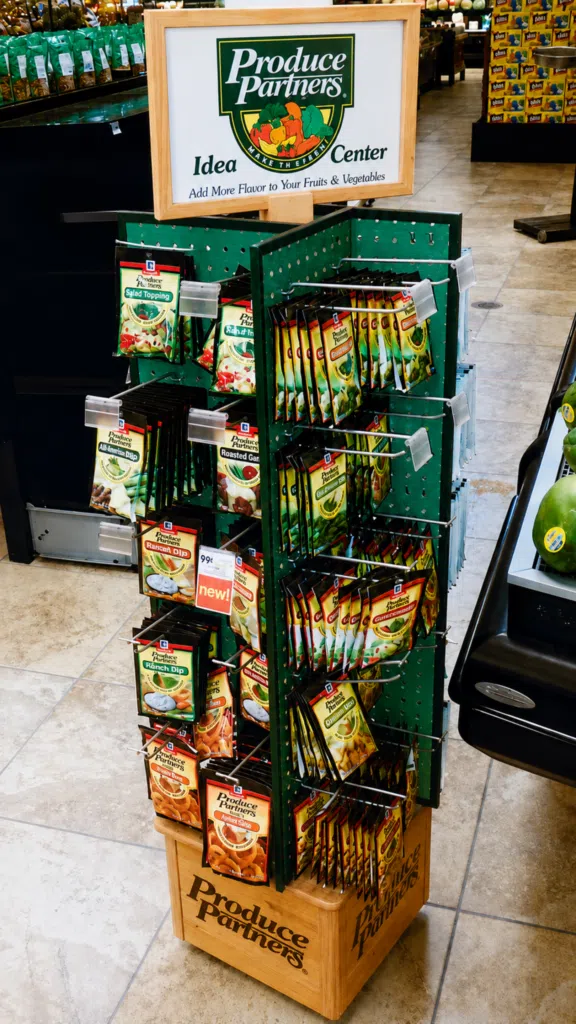 Peg Hook Display | BAW Plastics Inc. | BAW Plastics Inc. Display stand labeled 'Produce Partners' with packets of salad toppers and dressings on a green rack in a grocery store.