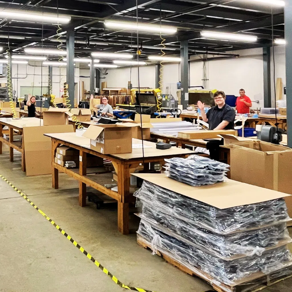 Factory workspace with workers at long benches handling cardboard boxes and stacks of material under bright fluorescent lights, yellow cords hanging from the ceiling.